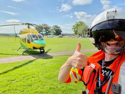 A thumbs up selfie of Dr Reuben Cooper wearing a flight helmet and visor with the Wiltshire Air Ambulance in the background. 