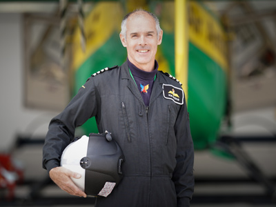 Pilot Rob in black flight suit, holding flight helmet with Wiltshire Air Ambulance's helicopter in the background