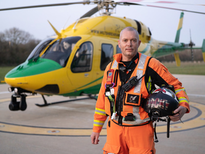 Paramedic Paul Rock holding flight helmet standing in front of Wiltshire Air Ambulance helicopter