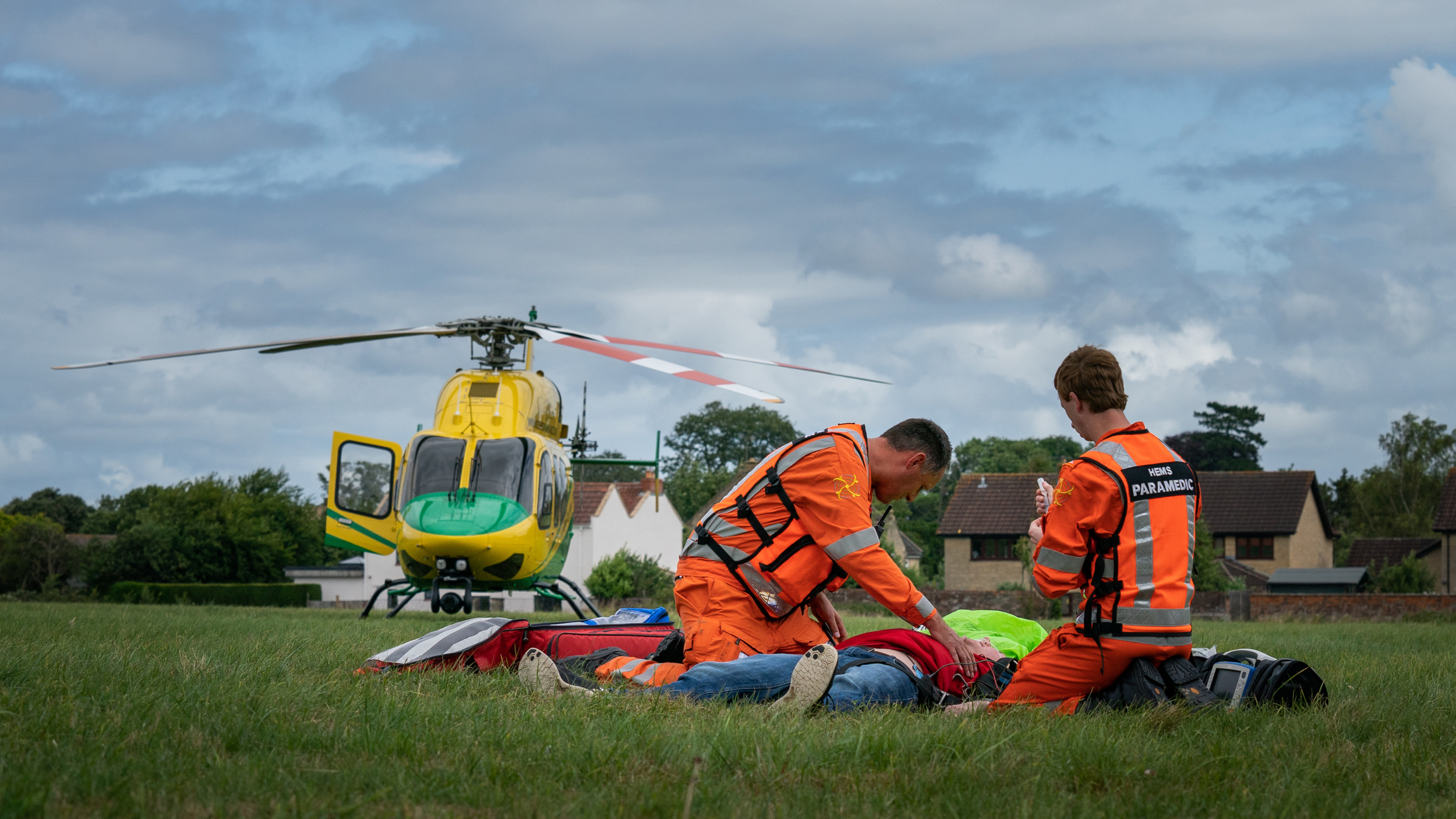 Two paramedics knelt next to a staged incident patient in a field with the helicopter in the background