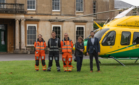 Two crew wearing orange, a pilot and two adults standing in front of a yellow and green helicopter in front of Hartham Park house