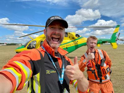 Dr Reuben Cooper and critical care paramedic Craig Wilkins taking a thumbs up selfie in a sunny field in front of the Wiltshire Air Ambulance helicopter.