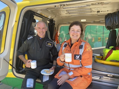 Pilot Rob and paramedic Jo posing with cakes and mugs whilst sat in Wiltshire Air Ambulance's helicopter