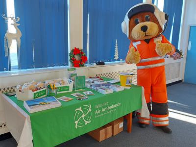 Paramedic bear mascot Wilber with his thumbs up, next to a table with Wiltshire Air Ambulance green table cloth and merchandise